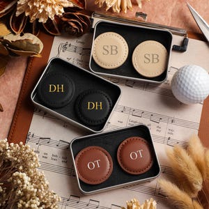 May include: An overhead shot features personalized golf ball markers in small metal tins. The markers are in black, tan, and brown leather, each with initials. A white golf ball and scissors are also visible.