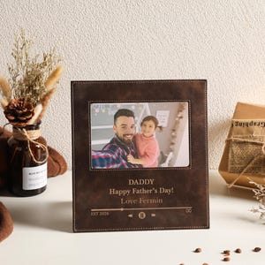 Puede incluir: Un marco de fotos rectangular de cuero marrón oscuro con costuras blancas. El marco presenta un mensaje grabado que dice: DADDY, Happy Father's Day!, Love Fermin, EST 2026, junto con un icono de interfaz de reproductor de música con pausa.