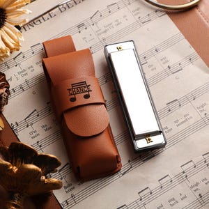 Puede incluir: Un estuche de armónica de cuero marrón con la palabra "CHANCE" y un símbolo de nota musical, junto a una armónica plateada. Los artículos descansan sobre una partitura con flores secas en el fondo.
