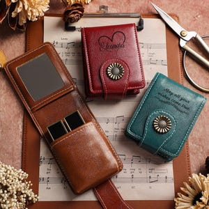 May include: A brown leather case with a mirror and black cufflinks, alongside two closed leather cases in red and teal. The red case is engraved with a heart and the name "Eleanora". The teal case has the text "May all your wishes come true".