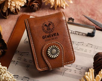 May include: A brown leather wallet with white stitching and a decorative metal clasp. The wallet is embossed with the name "GENEVRA" and the date "2025-10-23". The wallet is resting on sheet music.