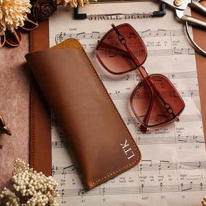 Puede incluir: Funda de gafas de sol de cuero marrón con las iniciales "LTK" en blanco, junto a unas gafas de sol cuadradas con un tinte marrón rojizo. Las gafas de sol descansan sobre una partitura con las palabras "Jingle Bells".