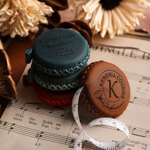 Può includere: Una pila di metri a nastro personalizzati in pelle verde acqua, verde e rossa. Uno è inciso con "Clara's tape measure", un altro con "Kendall Cousins" e un motivo floreale. È visibile un metro a nastro bianco.