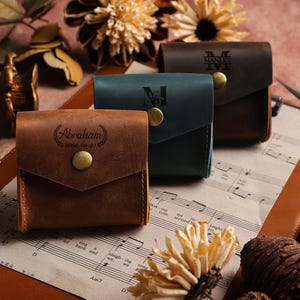 Puede incluir: Tres carteras de cuero en marrón, verde azulado y marrón oscuro, cada una con solapa y botón de latón. La cartera marrón está grabada con el nombre "Abraham". La cartera marrón oscuro está grabada con el nombre "Manley".