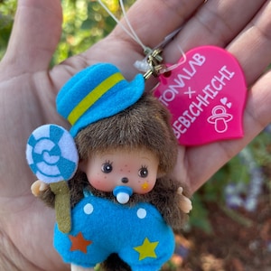 Puede incluir: Una muñeca Monchhichi con un sombrero azul con una raya amarilla, un mono azul con detalles de estrellas y sosteniendo una piruleta azul y blanca. La muñeca tiene un chupete y una etiqueta rosa que dice "TONALAB x BEBICHHICHI".