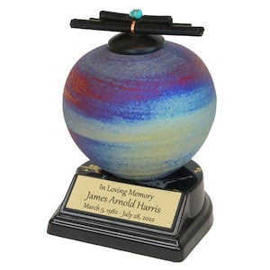 Op de afbeelding: Een blauwe, rode en gouden keramische urn met een zwarte basis en een houten bovenkant. De urn heeft een gepersonaliseerde plaquette met de tekst "In Loving Memory James Arnold Harris 5 maart 1982 - 28 juli 2022".