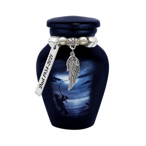 May include: Navy blue cremation urn with a fishing scene and a silver charm. The urn has a dark blue lid and a decorative band with pearls. A silver tag reads "Dad 1954-2021". The charm features a wing and a rose.