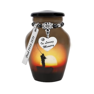 May include: Brown cremation urn with a black lid, featuring a sunset scene with a fisherman. It has a silver charm that says "In Loving Memory" and a tag that says "Dad 1954-2021". The urn is designed to hold the ashes of a loved one.