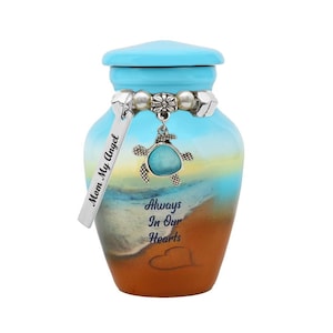 May include: A small blue and brown ceramic urn with a sea turtle charm and a silver tag that reads "Mom My Angel". The urn has a beach scene with the words "Always In Our Hearts" on the front.