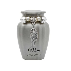 May include: Silver cremation urn with a white pearl and silver charm necklace with a rose and angel wing pendant. The urn is engraved with "Mom 1956-2024".