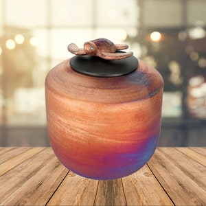 May include: A decorative urn with a turtle figurine on the lid. The urn has a rounded shape and a color gradient from orange to blue. The turtle is bronze-colored with a blue gem eye. The urn is sitting on a wooden surface.