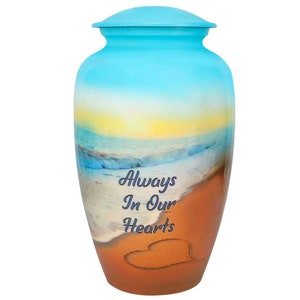 May include: A blue and orange cremation urn with a beach scene and the words "Always In Our Hearts" on the front. A heart is drawn in the sand.