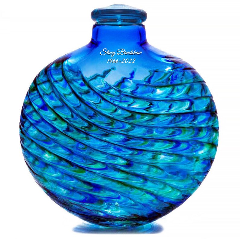 May include: A blue glass urn with a swirling pattern. The urn has a round shape and a small opening at the top. The urn is inscribed with the name "Stacy Bradshaw" and the dates "1966-2022".