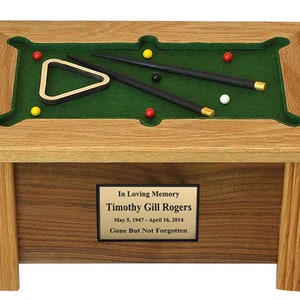 May include: A wooden pool table shaped cremation urn with a green felt top, two black pool cues, a triangle rack, and six billiard balls. The urn has a brass plaque with the inscription "In Loving Memory Timothy Gill Rogers May 5, 1947 - April 16, 2014 Gone But Not Forgotten."