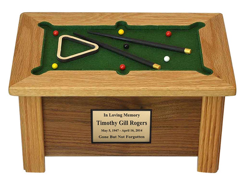 May include: A wooden pool table shaped cremation urn with a green felt top, two black pool cues, a triangle rack, and six billiard balls. The urn has a brass plaque with the inscription "In Loving Memory Timothy Gill Rogers May 5, 1947 - April 16, 2014 Gone But Not Forgotten."