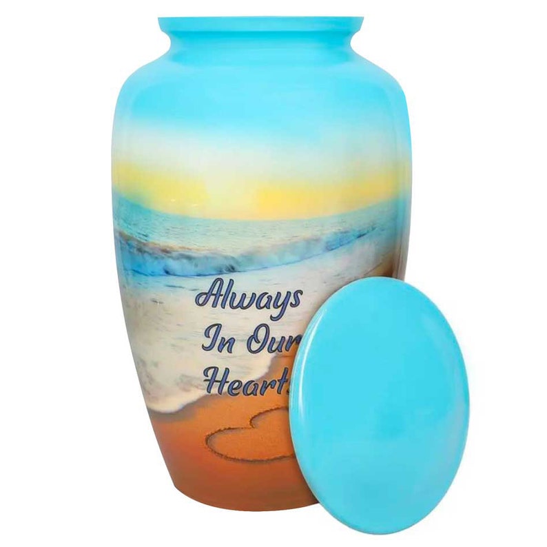 May include: A blue and brown cremation urn with a beach scene and the words "Always In Our Hearts" on the front. The urn has a heart drawn in the sand.