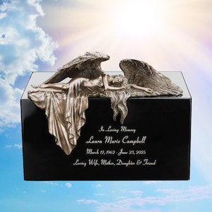 May include: A black memorial urn with a bronze angel sculpture. The angel is lying down with outstretched wings. The urn has the inscription "In Loving Memory" and the name "Laura Marie Campbell" with dates.