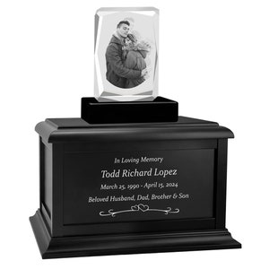 May include: Black wooden urn with a crystal photo memorial on top. The crystal photo shows a couple embracing. The urn has an engraved inscription that reads "In Loving Memory Todd Richard Lopez March 25, 1990 - April 15, 2024 Beloved Husband, Dad, Brother & Son".