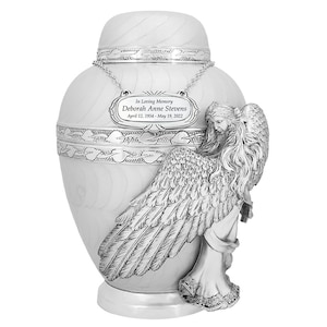 May include: White ceramic urn with a silver angel figurine. The urn has a silver plaque with the text "In Loving Memory Deborah Anne Stevens April 12, 1954 - May 19, 2022".
