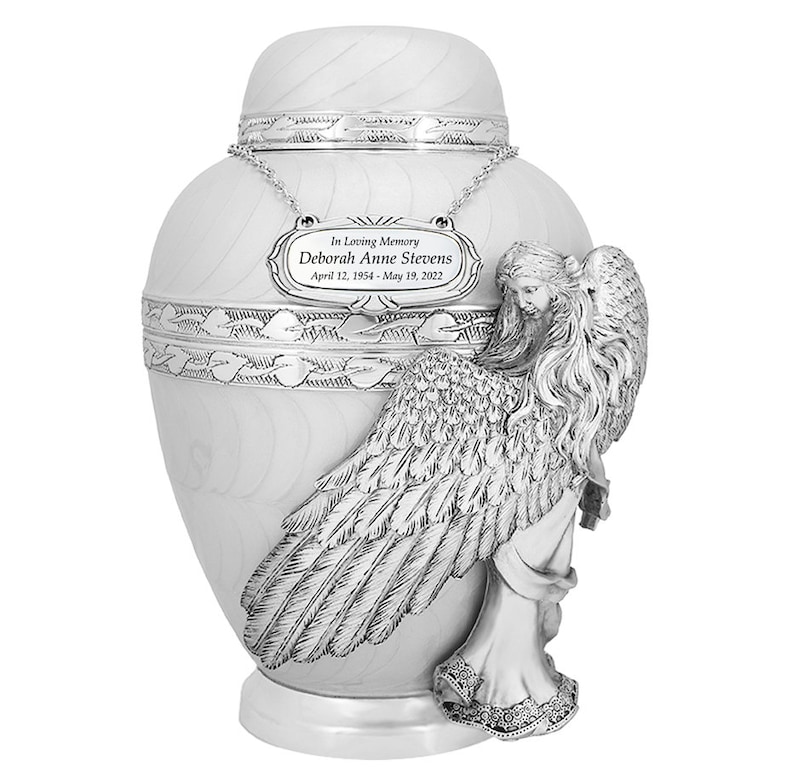 May include: White ceramic urn with a silver angel figurine. The urn has a silver plaque with the text "In Loving Memory Deborah Anne Stevens April 12, 1954 - May 19, 2022".