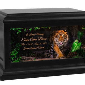 May include: Black wooden cremation urn with a photo of a tiger walking through a jungle. The urn has a personalized inscription that reads "In Loving Memory Elaine Annie Blaine Nov. 7, 1958 - Aug. 16, 2021 Forever Loved & Missed"