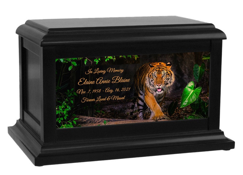 May include: Black wooden cremation urn with a photo of a tiger walking through a jungle. The urn has a personalized inscription that reads "In Loving Memory Elaine Annie Blaine Nov. 7, 1958 - Aug. 16, 2021 Forever Loved & Missed"