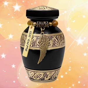 May include: Black cremation urn with gold accents and an engraved angel wing charm. The urn has a personalized tag that reads "Dad 1951-2025".