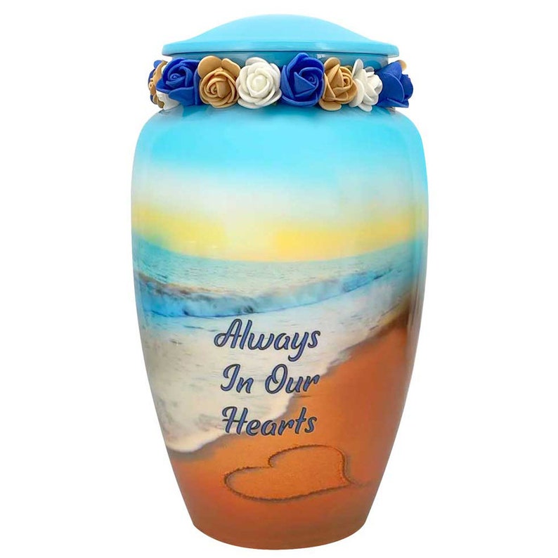 May include: A blue and orange ceramic urn with a beach scene and the words "Always In Our Hearts" on the front. The urn is decorated with a heart drawn in the sand and a floral wreath around the lid.