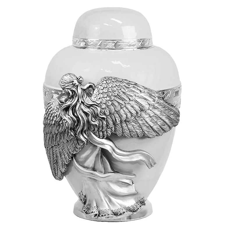 May include: White ceramic urn with a silver angel sculpture on the side. The angel has wings and flowing hair. The urn has a decorative band around the top.
