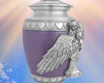 Puede incluir: Una urna funeraria lavanda con tapa y base plateadas, adornada con recortes en forma de corazón. Una detallada escultura de ángel plateado está adherida al costado. La urna está diseñada para contener las cenizas de un ser querido.