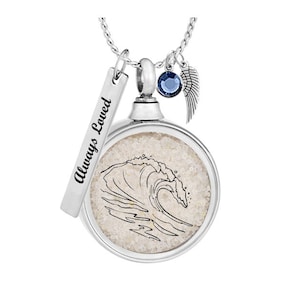 May include: Silver necklace with a round pendant containing white sand and a black line drawing of a wave. The pendant is attached to a silver chain with a small silver charm that says "Always Loved" and a blue crystal charm.