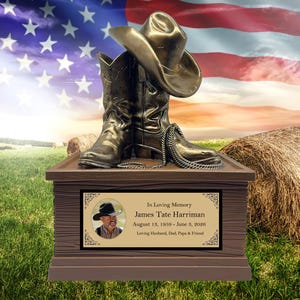 May include: A bronze-colored cowboy hat and boots rest on a wooden memorial box. The box features a photo and inscription: "In Loving Memory James Tate Harriman August 15, 1959 - June 3, 2026 Loving Husband, Dad, Papa & Friend."
