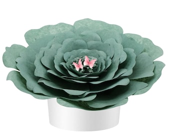 May include: A large, layered paper flower in shades of teal, with delicate pink butterflies nestled in the center. The flower sits in a white, cylindrical pot. This decorative piece could be used as a centerpiece or home decor item.