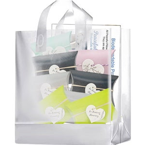 May include: A clear plastic bag with a white handle, filled with small, colorful, heart-shaped pillow boxes. The boxes have the text "In Loving Memory" printed on them. The bag is labeled "Biodegradable Products" and "Peaceful Pillow".