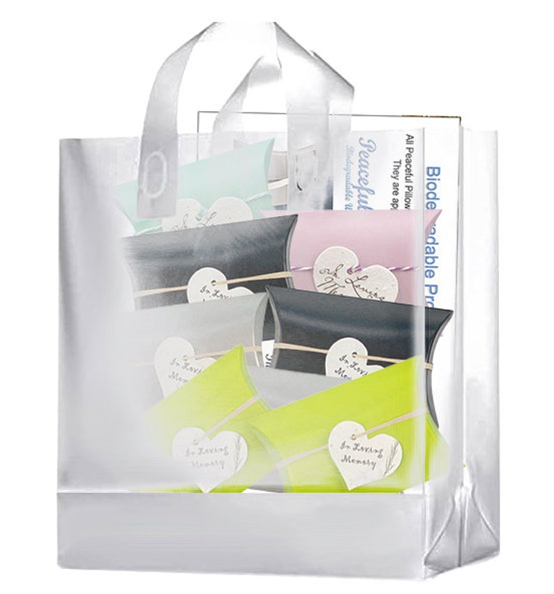 May include: A clear plastic bag with a white handle, filled with small, colorful, heart-shaped pillow boxes. The boxes have the text "In Loving Memory" printed on them. The bag is labeled "Biodegradable Products" and "Peaceful Pillow".