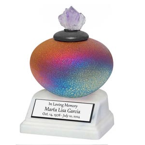 May include: A memorial urn with a color-shifting, spherical body, transitioning from orange to blue. It features a black base, a white pedestal, and a purple crystal on top. The urn includes a plaque with the inscription "In Loving Memory Marta Lisa Garcia Oct. 14, 1978 - July 10, 2024."