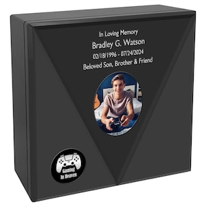 May include: A black wooden box with a photo of a young man holding a video game controller. The box has a silver inscription that reads "In Loving Memory Bradley G. Watson 02/18/1996 - 07/24/2024 Beloved Son, Brother & Friend". The box also has a black and white icon of a video game controller with the text "Gaming In Heaven".