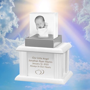 May include: A white memorial urn with a rectangular, clear glass block on top, displaying a black and white photo of a sleeping baby. The urn has a decorative front panel with text: "Our Little Angel Jonathan Ryan Martin January 12, 2025 Always In Our Hearts."