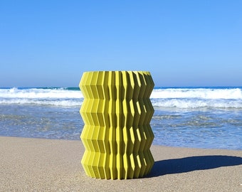 May include: A yellow geometric patterned cylinder vase on a sandy beach with blue ocean waves in the background.