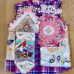 Può includere: Un gilet patchwork con un motivo a quadri viola e rosa. Il gilet è adornato con varie toppe di tessuto, tra cui pizzo, centrini e dettagli ricamati. Una grafica di uccelli e farfalle è presente su una delle toppe. Bottoni ed elementi decorativi sono cuciti sul gilet.