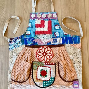 May include: A patchwork apron with a variety of fabrics, including floral, polka dot, and patterned textiles. The apron features a blue top with a red and white square design, a large pocket, and lace trim. The apron has a white neck strap and waist ties.