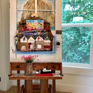 May include: A brown plaid button-down shirt featuring a colorful fabric collage of houses and a bird, displayed on a wooden easel. Below the shirt, a brown vase with pink flowers and a small black and white toy boat sit on the easel shelf.