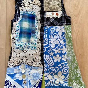 May include: A sleeveless waistcoat constructed from a black and white plaid shirt base, featuring various fabric patches including blue floral prints, white eyelet lace, green patterned fabric, and a row of buttons on a blue floral panel.