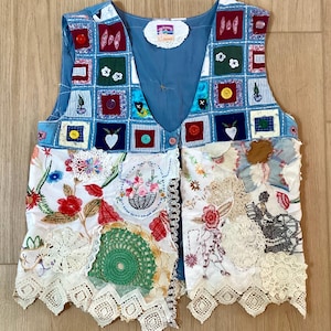 May include: A patchwork vest with a blue V-neck and button closure. The vest features a variety of fabric squares with embroidered designs, buttons, and lace trim. The vest is a mix of colours, including blue, red, green, and white.