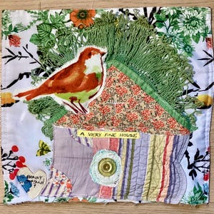 May include: A fabric art piece featuring a brown and white bird applique, a green fringe, and floral patterned fabrics. The text "A VERY FINE HOUSE" is visible. The piece is a patchwork of various fabrics.
