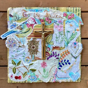May include: A mixed media artwork featuring a bird, a crucifix, and a clock. The artwork incorporates fabric scraps with floral and embroidered details. The text "LOVE LIFE To the Fullest" is visible. The piece has a vintage, handcrafted aesthetic.