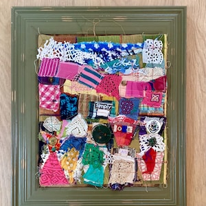 May include: A framed textile collage featuring a grid of multicolored fabric scraps, lace, and buttons. The composition includes various patterns, textures, and a small label with the word Simply. The entire piece is encased in a green frame.