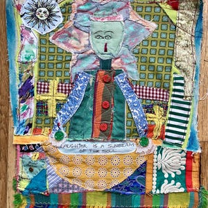 May include: A colorful patchwork quilt with a whimsical figure in the center. The figure has a green face, a red nose, and is wearing a green jacket with red buttons. The quilt is made of various fabrics and textures, including plaid, gingham, and floral patterns. The quilt has a fringe border and the text "Laughter is a sunbeam of the soul" is stitched on the bottom.