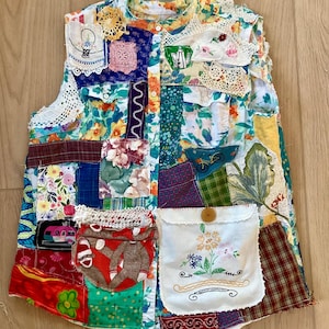 May include: A patchwork vest featuring a variety of fabric patterns and textures. The vest includes floral prints, lace, and embroidered details. A pocket with floral embroidery and a wooden button closure is visible. The vest is a unique, handmade garment.