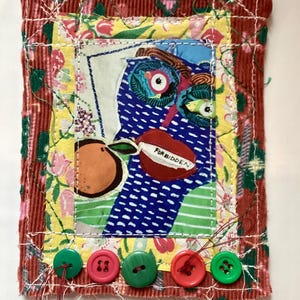May include: A rectangular fabric collage featuring a central blue patterned figure with large eyes and red lips holding a peach. The word FORBIDDEN is stitched on the lips. The piece is bordered by floral yellow and red corduroy fabric with five buttons.